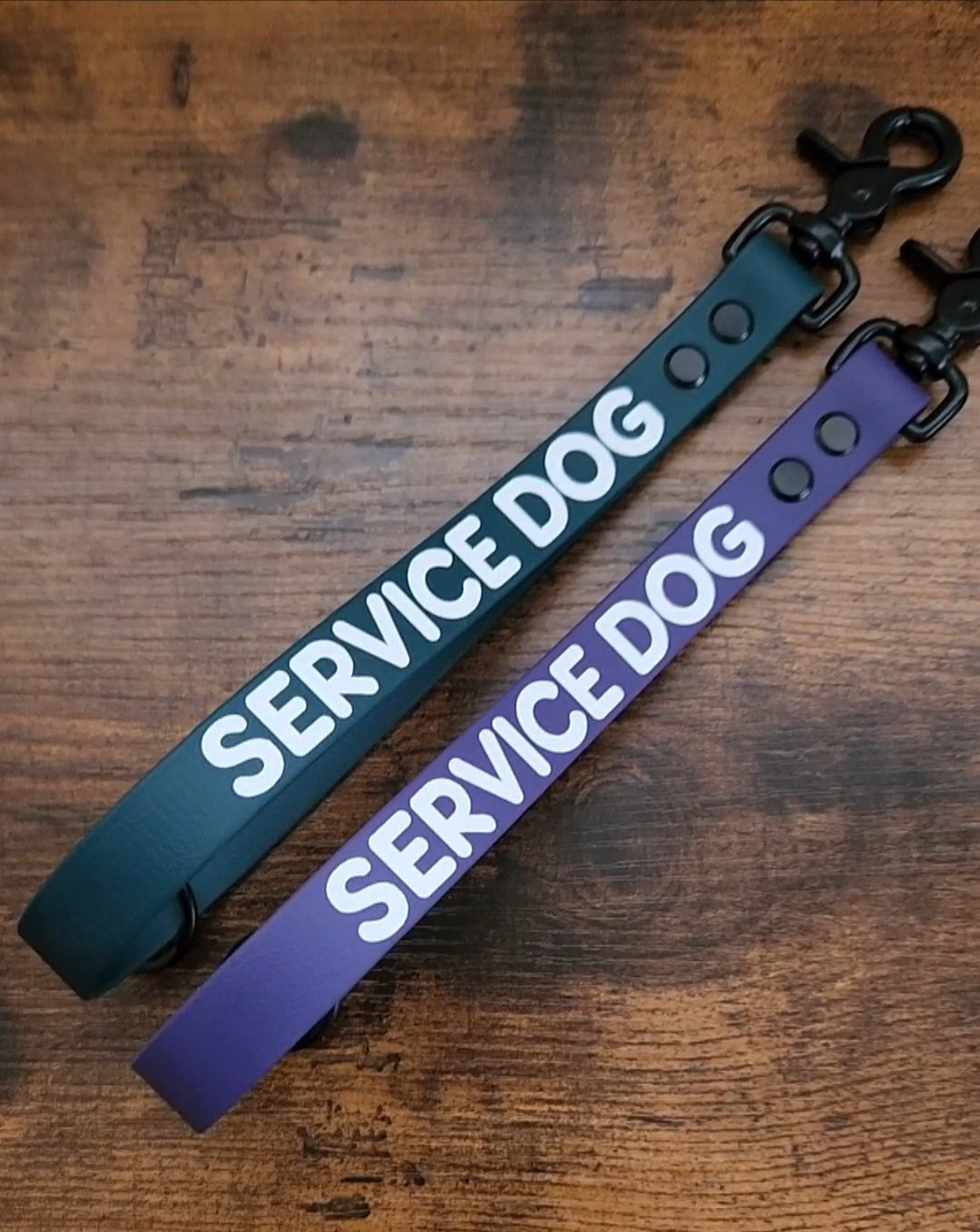 two traffic handles with white text reading "SERVICE DOG" one on dark green biothane and one on dark purple biothane