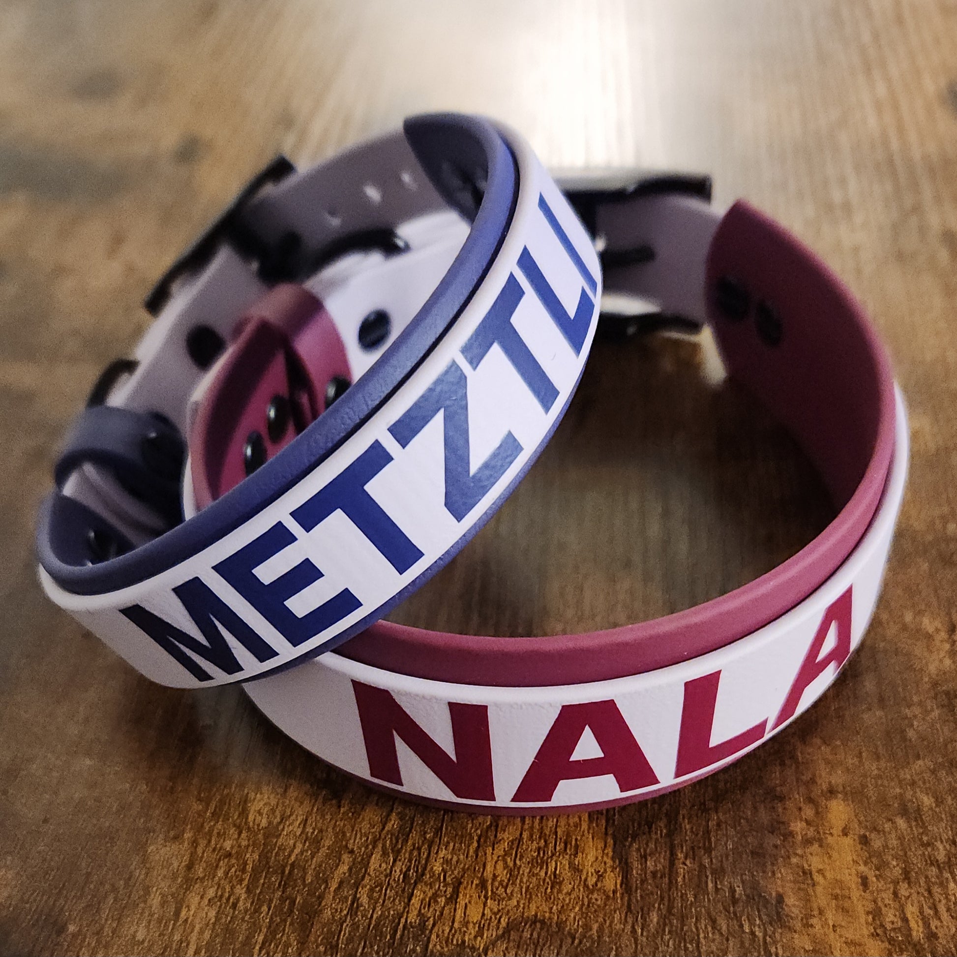 two dog collars, one light and dark purple layered biothane reading "METZTLI" and the other wine and light purple layered biothane reading "NALA"